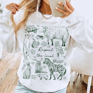 May include: White sweatshirt with a green illustration of various animals, including a zebra, owl, and turtle. The text "Respect the local" is printed in green. The sweatshirt has a crew neck and long sleeves.
