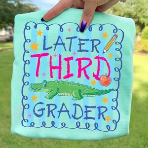 Op de afbeelding: Lichtblauw t-shirt met de woorden "Later Third Grader" in roze en blauw. Het ontwerp bevat een groene alligator, een potlood, een appel en sterren. Het shirt wordt door een hand omhoog gehouden.