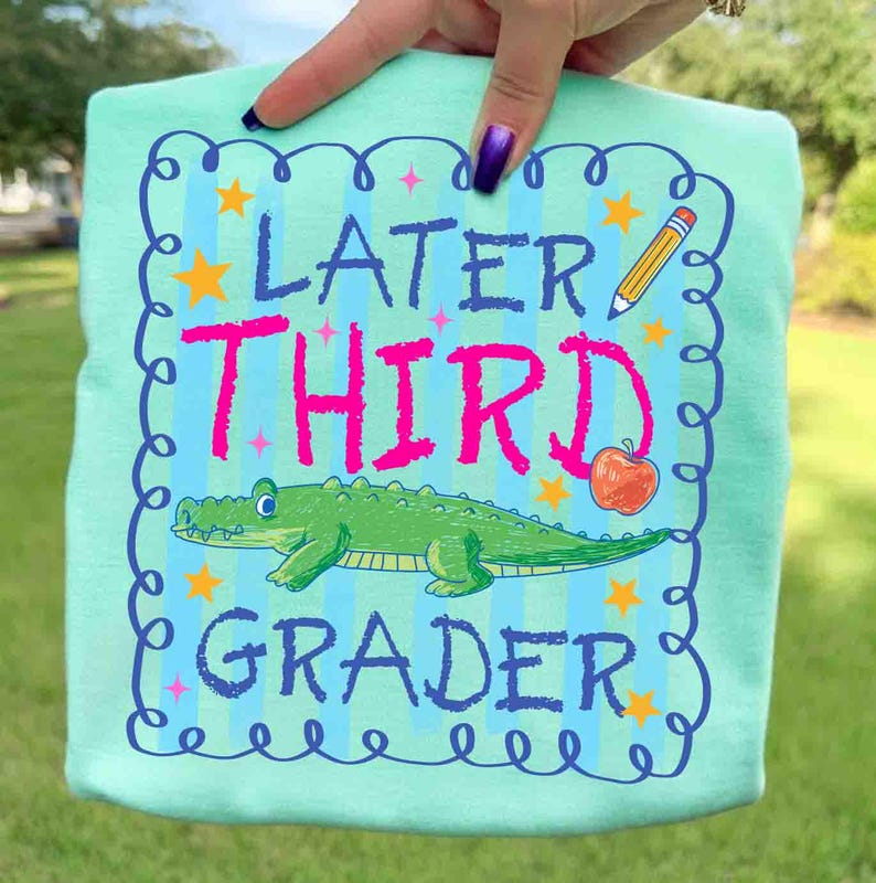 Op de afbeelding: Lichtblauw t-shirt met de woorden "Later Third Grader" in roze en blauw. Het ontwerp bevat een groene alligator, een potlood, een appel en sterren. Het shirt wordt door een hand omhoog gehouden.