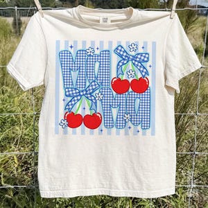 May include: Cream-colored t-shirt with a graphic design featuring the word "MOM" in blue gingham letters. Red cherries and blue bows accent the design, with white flowers and blue stripes completing the look. The shirt is hanging on a clothesline.
