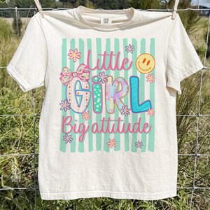 May include: A cream-coloured short-sleeve t-shirt featuring a graphic print with vertical teal stripes. The text reads Little Girl Big attitude in pink and blue. The design includes a patterned bow, small flowers, and a yellow smiley face icon.