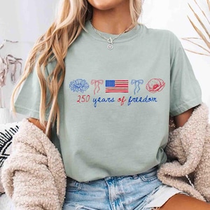 May include: A light green t-shirt with a patriotic design. The design includes a blue flower, red bows, an American flag, and a red poppy. The text "250 years of freedom" is written in blue script. The shirt is a casual style.