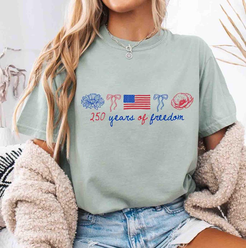 May include: A light green t-shirt with a patriotic design. The design includes a blue flower, red bows, an American flag, and a red poppy. The text "250 years of freedom" is written in blue script. The shirt is a casual style.