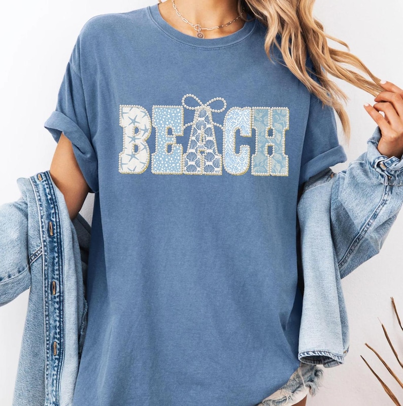May include: A blue t-shirt featuring the word "BEACH" in a decorative font. The letters are filled with blue and white patterns, including starfish and seashell designs. The shirt has rolled-up sleeves and a relaxed fit.