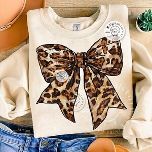 May include: A beige sweatshirt with a large leopard print bow design. The bow is drawn in a realistic style with brown and black spots.