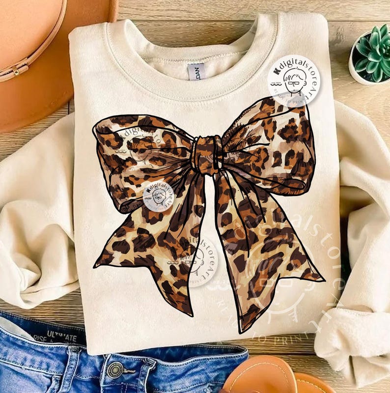 May include: A beige sweatshirt with a large leopard print bow design. The bow is drawn in a realistic style with brown and black spots.