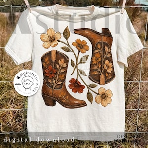 May include: Cream-colored t-shirt featuring a graphic of brown cowboy boots and floral accents. The design includes the words "digital download" and "designed by studio". The shirt is hanging on a clothesline.