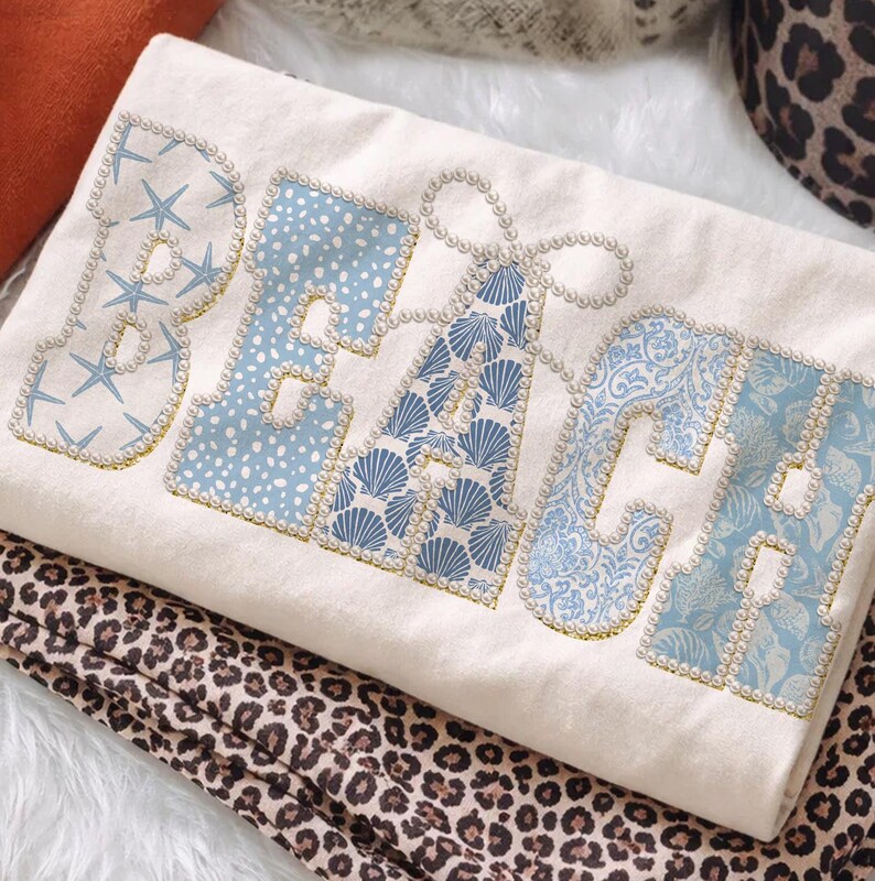 May include: Cream-coloured decorative pillow with the word "BEACH" in blue and white patterned letters. The letters are outlined with faux pearls and gold trim. The pillow is resting on a leopard print fabric.