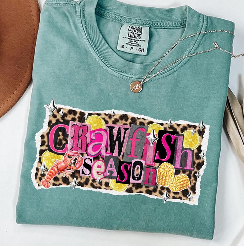 May include: A teal Comfort Colors t-shirt with the words "Crawfish Season" in a collage-style design. The design includes a crawfish, lemons, and corn on a leopard print background. A gold necklace with a pendant is draped on the shirt.