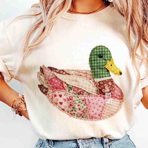 May include: Cream-colored t-shirt featuring a patchwork duck design. The duck has a green checkered head, a yellow beak, and a body composed of various pink and floral patterned fabrics. The text "DO NOT COPY" is visible on the duck's body.