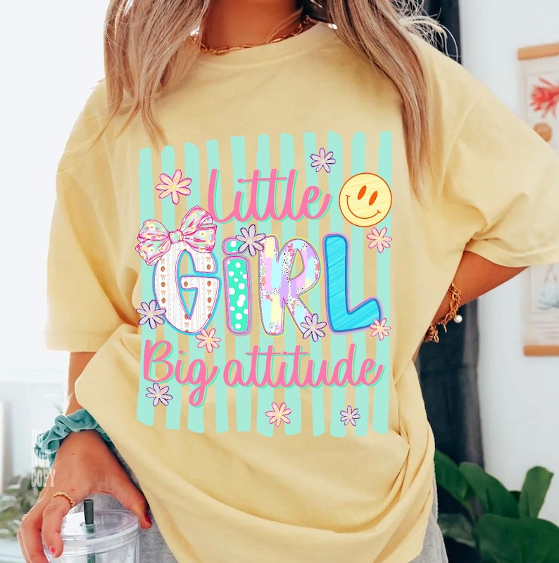 May include: A pale yellow t-shirt featuring a graphic print with vertical light blue stripes. The text reads Little Girl Big attitude in pink and blue. The design includes a patterned bow, small flowers, and a yellow smiley face icon.