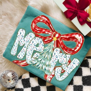May include: Teal Christmas pillow with a red and white plaid bow and the word "MERRY" in white script with a blue and green leaf pattern. A small Christmas tree is in the center of the bow.
