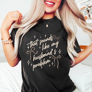 May include: Black t-shirt with the phrase "that sounds like my husband's problem" in a cursive font, accented with bow graphics and stars. The shirt is a dark color and has a crew neck.