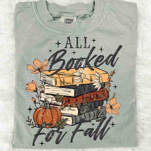 May include: Sage green t-shirt with a graphic of stacked books, a pumpkin, and flowers. The text reads "ALL Booked for Fall" in a decorative font. The shirt is made of a soft material.