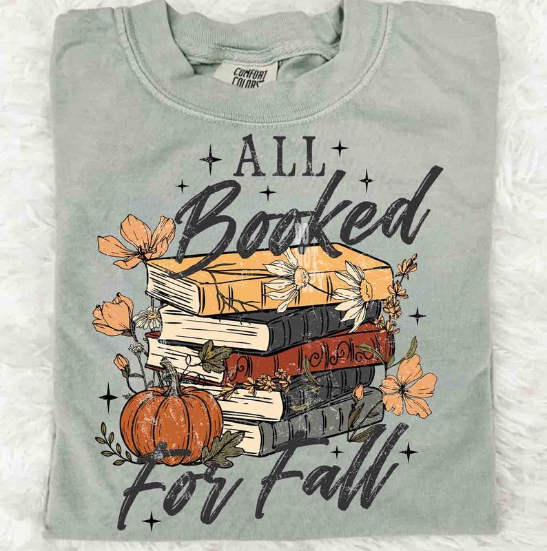 May include: Sage green t-shirt with a graphic of stacked books, a pumpkin, and flowers. The text reads "ALL Booked for Fall" in a decorative font. The shirt is made of a soft material.