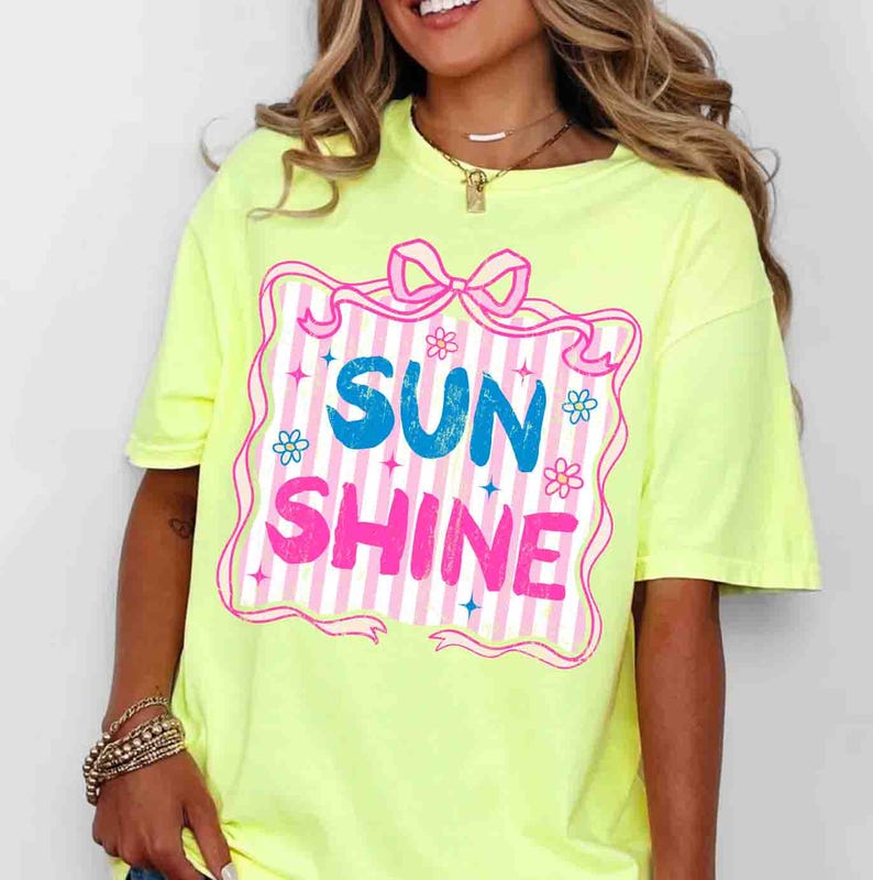May include: A neon yellow t-shirt featuring the words "SUN SHINE" in blue and pink, framed by a pink border with a bow and floral accents. The shirt has short sleeves and a crew neck.