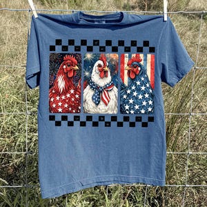 May include: Blue t-shirt featuring three patriotic chicken portraits. Each chicken is decorated with red, white, and blue patterns, including stars, stripes, and fireworks. A black and white checkered border frames the design. The shirt is hanging on a clothesline.