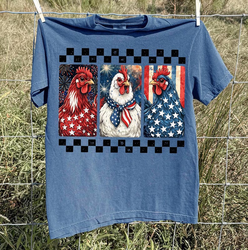 May include: Blue t-shirt featuring three patriotic chicken portraits. Each chicken is decorated with red, white, and blue patterns, including stars, stripes, and fireworks. A black and white checkered border frames the design. The shirt is hanging on a clothesline.