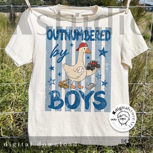 May include: Cream-colored t-shirt with a blue and white striped background. The shirt features a cartoon goose wearing a red cap and holding a baseball and a monster truck. Text reads "OUTNUMBERED by BOYS."