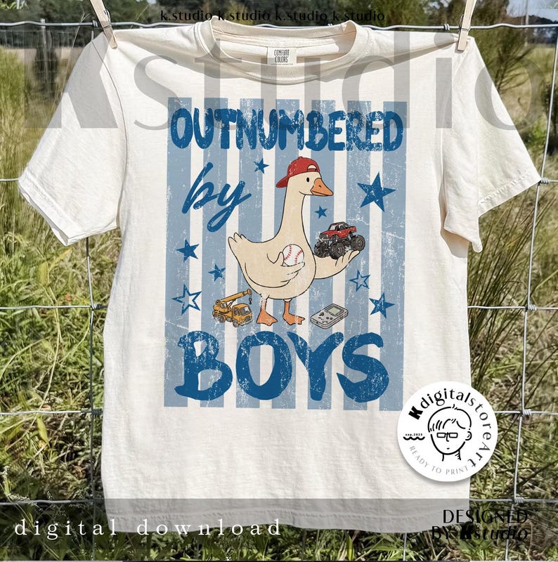 May include: Cream-colored t-shirt with a blue and white striped background. The shirt features a cartoon goose wearing a red cap and holding a baseball and a monster truck. Text reads "OUTNUMBERED by BOYS."