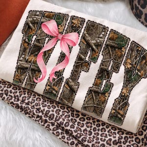 Puede incluir: Sudadera color crema con la palabra "Wifey" en un patr&oacute;n de camuflaje, acentuada con un lazo rosa. La sudadera est&aacute; doblada sobre una tela con estampado de leopardo, creando un efecto de capas.