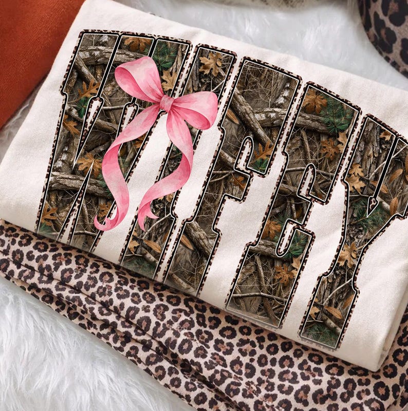 Puede incluir: Sudadera color crema con la palabra "Wifey" en un patr&oacute;n de camuflaje, acentuada con un lazo rosa. La sudadera est&aacute; doblada sobre una tela con estampado de leopardo, creando un efecto de capas.