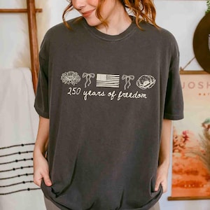 May include: Dark gray t-shirt with a white graphic. The graphic includes a flower, bows, an American flag, and a poppy, with the text "250 years of freedom" in a script font. The shirt has a crew neck and short sleeves.
