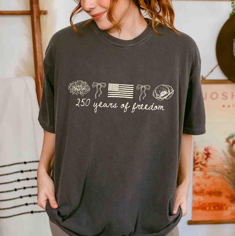 May include: Dark gray t-shirt with a white graphic. The graphic includes a flower, bows, an American flag, and a poppy, with the text "250 years of freedom" in a script font. The shirt has a crew neck and short sleeves.
