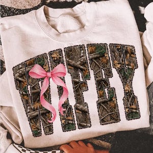 Puede incluir: Sudadera color crema con la palabra "WIFEY" en letras grandes. Las letras est&aacute;n rellenas con un patr&oacute;n de camuflaje y delineadas con un estampado de leopardo. Un lazo rosa est&aacute; centrado en la parte delantera.