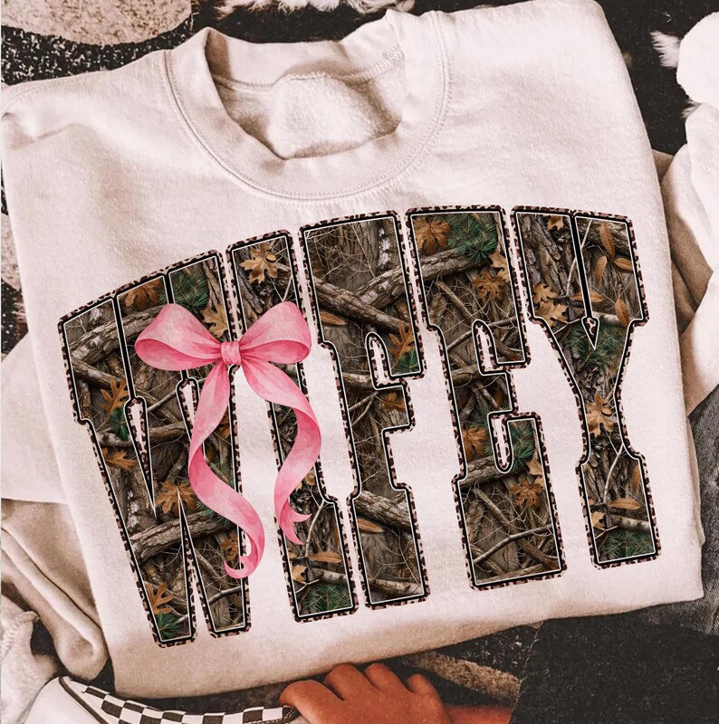 Puede incluir: Sudadera color crema con la palabra "WIFEY" en letras grandes. Las letras est&aacute;n rellenas con un patr&oacute;n de camuflaje y delineadas con un estampado de leopardo. Un lazo rosa est&aacute; centrado en la parte delantera.