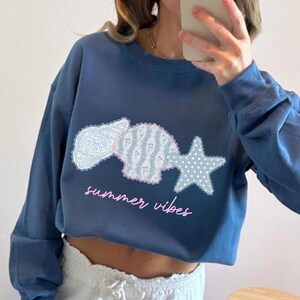May include: A blue cropped sweatshirt with a seashell, a shell, and a star graphic. The seashell and shell are patterned with floral and lace designs. The star is white with blue polka dots. The words "summer vibes" are written in pink script.