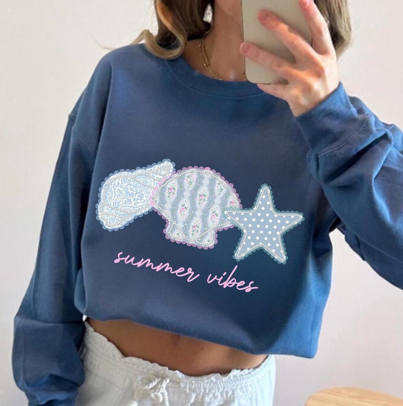 May include: A blue cropped sweatshirt with a seashell, a shell, and a star graphic. The seashell and shell are patterned with floral and lace designs. The star is white with blue polka dots. The words "summer vibes" are written in pink script.