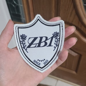 Może przedstawiać: Biała naszywka z tkaniny w kształcie tarczy z czarną obwódką. Naszywka ma czarne logo z napisem "ZBI" z tekstem "SINCE 2023" pod logo. Po obu stronach logo znajdują się dwie czarne stylizowane róże.