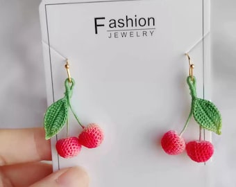 May include: Crocheted cherry earrings with green leaves and gold hooks. The earrings are handmade and have a pink and red gradient.