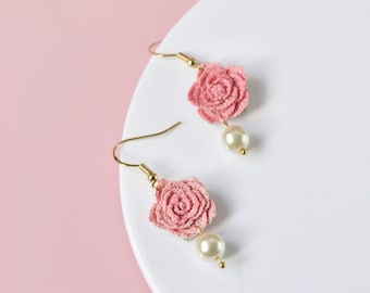 May include: A pair of delicate earrings featuring pink crocheted rose flowers with a pearl accent. The earrings have gold hooks.