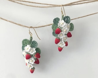 May include: A pair of crocheted strawberry earrings with white flowers and green leaves. The earrings are hanging from a string.