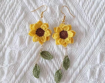 May include: Crocheted sunflower earrings with yellow petals and a brown center. The earrings have gold-colored hooks and dangling green leaves, creating a floral design. The earrings are displayed on a white textured surface.