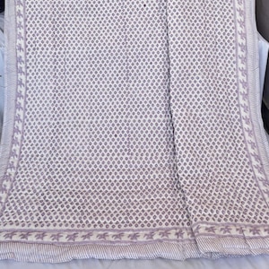May include: A white and lavender patterned quilt. The quilt has a repeating dot pattern in lavender across the main body. The edges are bordered with a lavender stripe and star design. The quilt is folded over a surface.