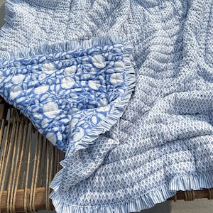 May include: A blue and white patterned quilt with a floral design and ruffled edges. The quilt is draped over a wooden chair. The quilt features a diamond pattern and a floral pattern. The quilt is made of a soft, quilted material.