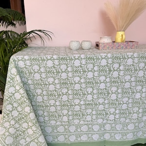 Puede incluir: Un mantel verde con un patrón floral blanco. El mantel está puesto sobre una mesa con dos pequeños candelabros blancos y una bandeja con una jarra amarilla y un tarro blanco encima.