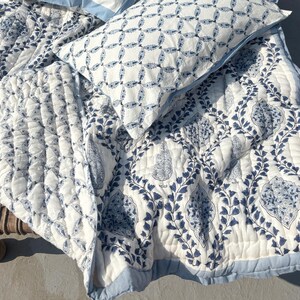 May include: A white and blue quilted bedding set with two matching pillows. The quilt and pillows feature a repeating floral pattern in shades of blue on a white background. The pillows have a blue border. The bedding is displayed on a wooden bench.