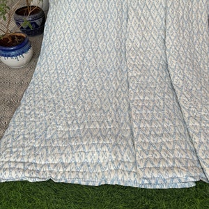 Könnte beinhalten: Eine weiße und hellblaue gemusterte Steppdecke mit einem Rautenmuster. Die Steppdecke ist gefaltet und liegt auf einer grünen Grasfläche.