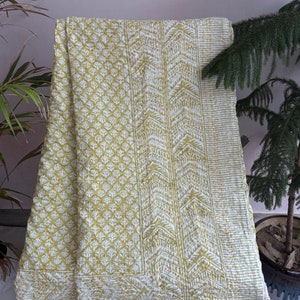May include: A yellow and white kantha quilt with a floral pattern and geometric designs. The quilt is folded and draped over a chair.