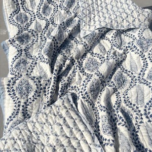 May include: A quilted blanket featuring a white base with a repeating blue botanical pattern of leaves and teardrop-shaped floral motifs. The fabric has a visible stitched texture throughout, with a solid light blue border along the edges.