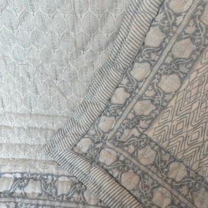 Puede incluir: Un textil acolchado con fondo blanco y un patrón repetitivo de hojas grises. El borde presenta una franja gris y blanca, un motivo de enredadera floral y un diseño geométrico de diamantes, todo en tonos de gris y blanco.