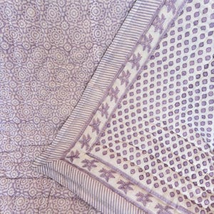 Peut inclure: Un textile matelassé présentant un motif géométrique violet clair et blanc sur la surface principale. La bordure affiche un fond blanc avec un motif floral violet répétitif et de fines rayures parallèles, créant un design de bord distinct.