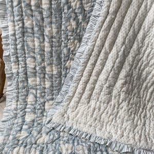 May include: A reversible quilted throw blanket featuring a blue and white floral pattern on one side and a small-scale geometric print on the other. The blanket is finished with a ruffled edge featuring a blue and white vertical stripe pattern.