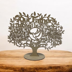 May include: A laser-cut wooden tree sculpture featuring the Lord's Prayer. The design includes a dove within a circle and the words of the prayer, with a tree trunk base. The sculpture is displayed on a wooden table.