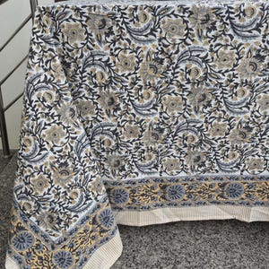 May include: A white tablecloth with a floral pattern in black, gray, and gold covers a table. The tablecloth has a border with a repeating floral design and a striped edge. The table is set on a gray surface.