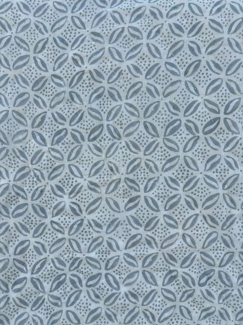 May include: A white fabric with a repeating pattern of blue stylized flowers. The flowers are made up of two overlapping teardrop shapes with a small cluster of dots in the center.